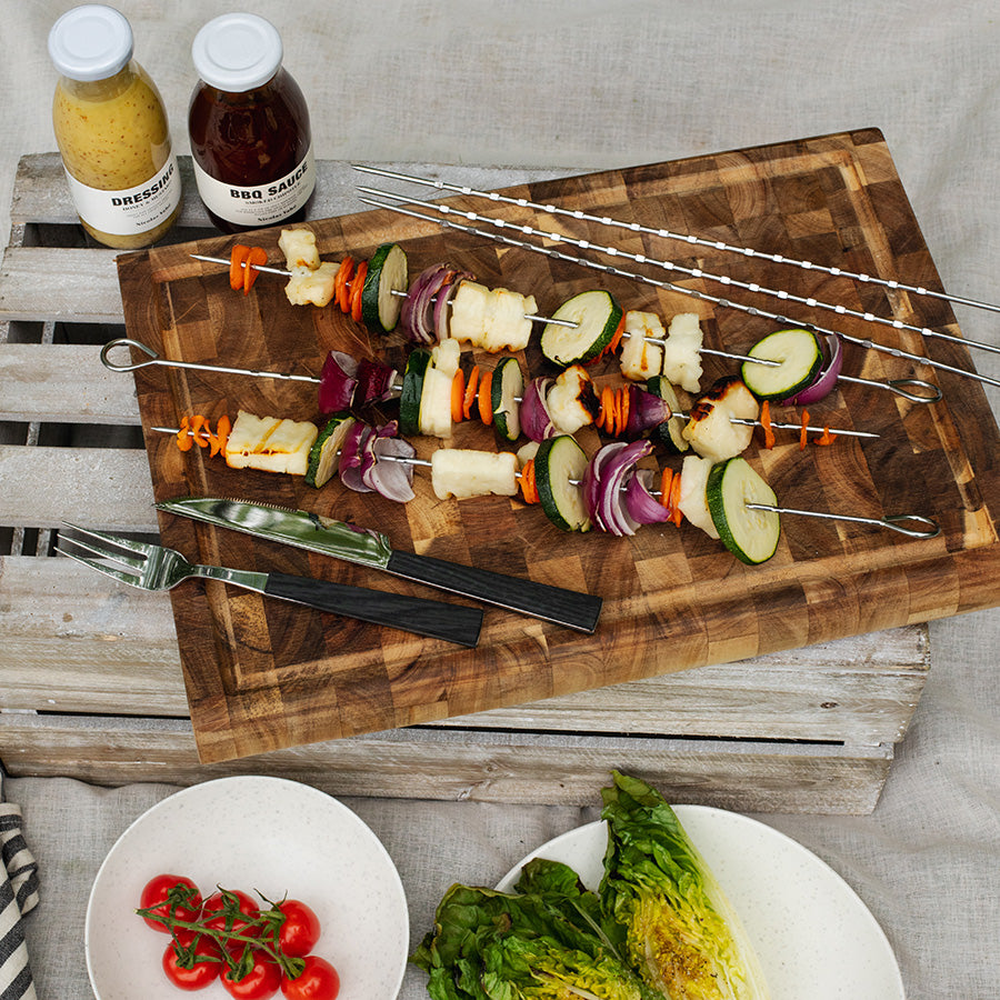 Seasons grillspyd med grilla grønnsaker og kjøtt på et skjærebrett, perfekt for grilling og servering. Nyttige tilbehør som BBQ-saus og dressing er synlige i bakgrunnen.