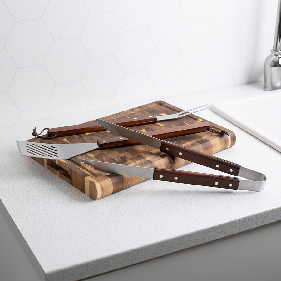 Seasons BBQ sett inkludert grilltang og stekespade liggende på et treskjærebrett, med et fokus på de robuste trehåndtakene og elegansen i børstet stål.