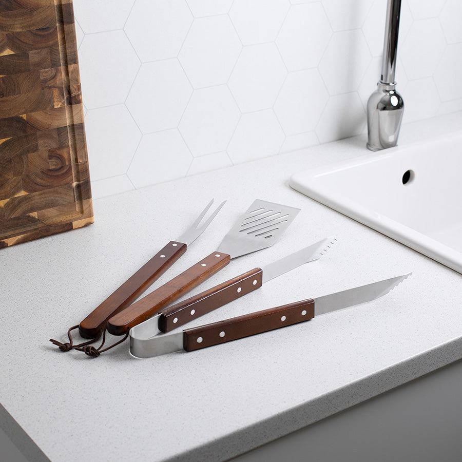 Seasons BBQ sett med grilltang, stekespade og grillgaffel i børstet stål med trehåndtak, plassert på en moderne kjøkkenbenk.
