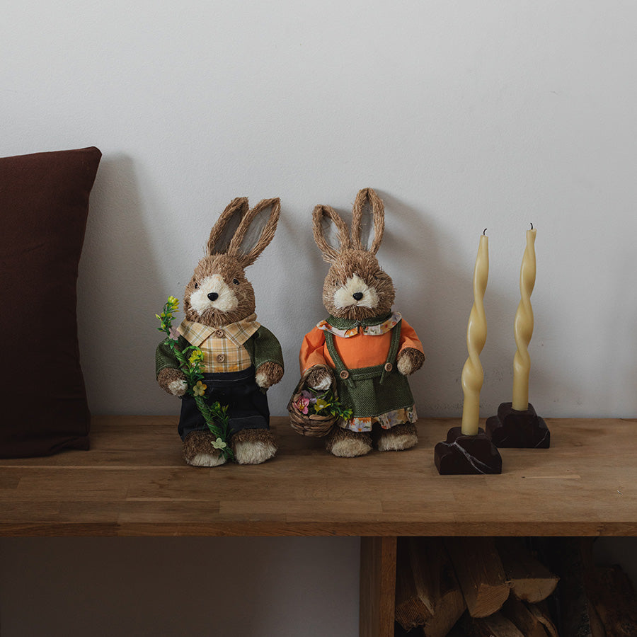 To haremor påskefigurer i halm står på en tregulv, med enkle detaljer som lyse lys og treverk i bakgrunnen, fra Seasons Easter Collection.