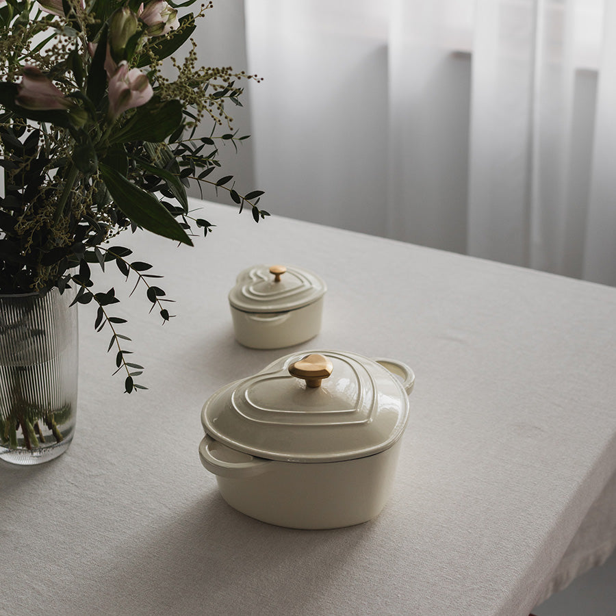 To hjerteformede støpejernsgryter i kremfarge, dekorativt plassert på et bord med blomster, ideelle for servering og dekor.