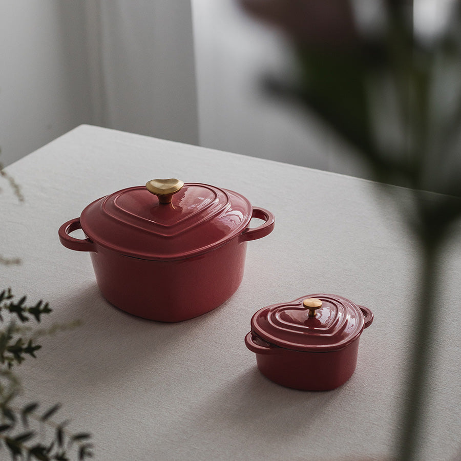 Støpejernsgryte i bringebærrød farge med gullknott, plassert ved siden av en mindre gryte på et lys bord.