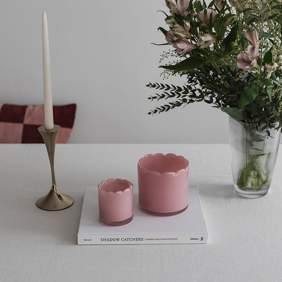 To lyslykter i rosa farget glass plassert på et bord, sammen med en elegant lysestake og blomster for en romantisk atmosfære.