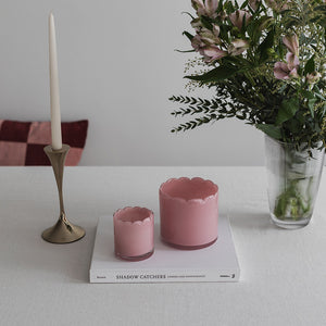 To lyslykter i rosa farget glass plassert på et bord, sammen med en elegant lysestake og blomster for en romantisk atmosfære.