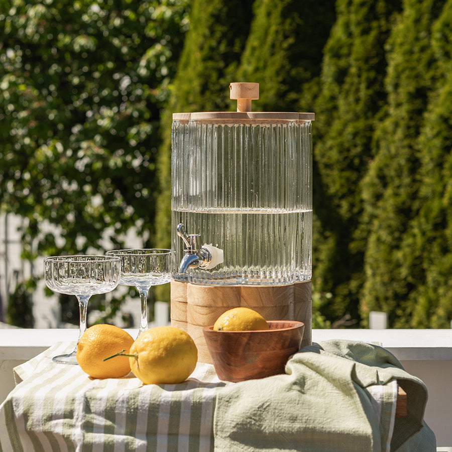 Drikkedispenser fra Seasons Summer Collection, glassbeholder med kran, plassert med sitroner og glass på en sommerlig duk, perfekt for hagefest og brunch.