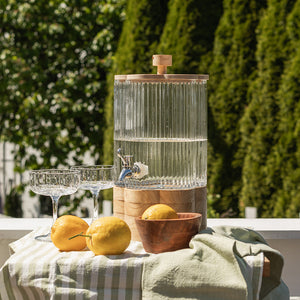 Drikkedispenser fra Seasons Summer Collection, glassbeholder med kran, plassert med sitroner og glass på en sommerlig duk, perfekt for hagefest og brunch.