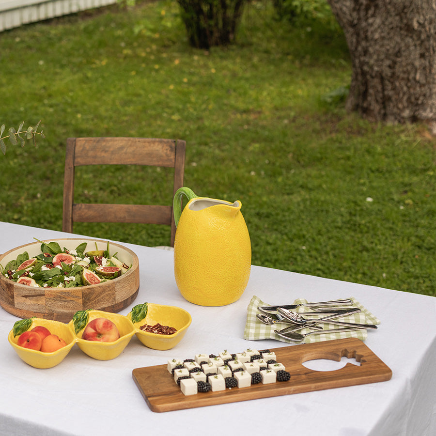 Fjøl i akasie på et bord i hagen, omringet av frisk salat, fruktboller og en gulterrakottakanne, skaper en innbydende sommerstund.