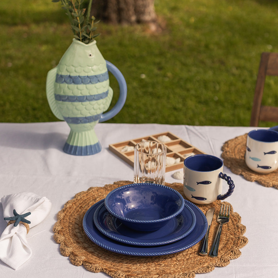 Et dekket bord med marineblå tallerken fra Seasons Summer Collection, omringet av en dekorativ fiskekanne og glass, ideelt for sommerlige måltider.