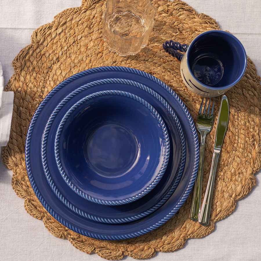 Marineblå tallerken i Seasons Summer Collection sett på en rustikk tallerkenunderlag, med matchende kopper og bestikk, perfekt for uteservering.