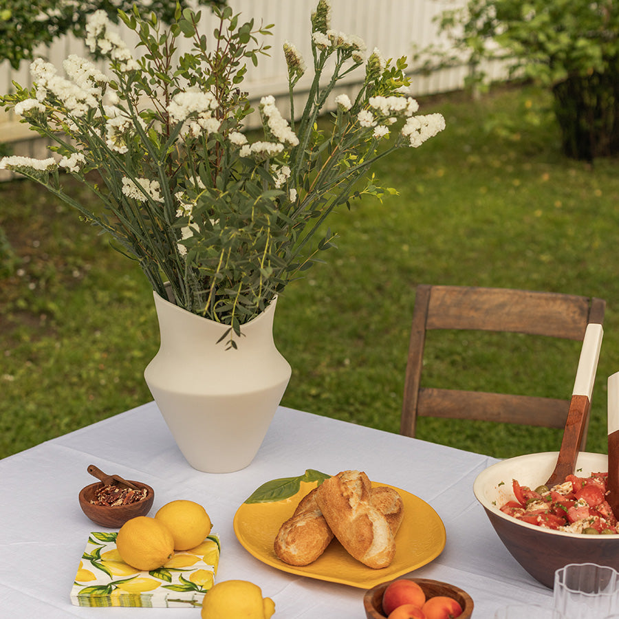 Stilig vase fra Seasons Summer Collection med små hvite blomster på et bord fylt med mat, sitroner og brød. Perfekt for sommerdekorasjon.