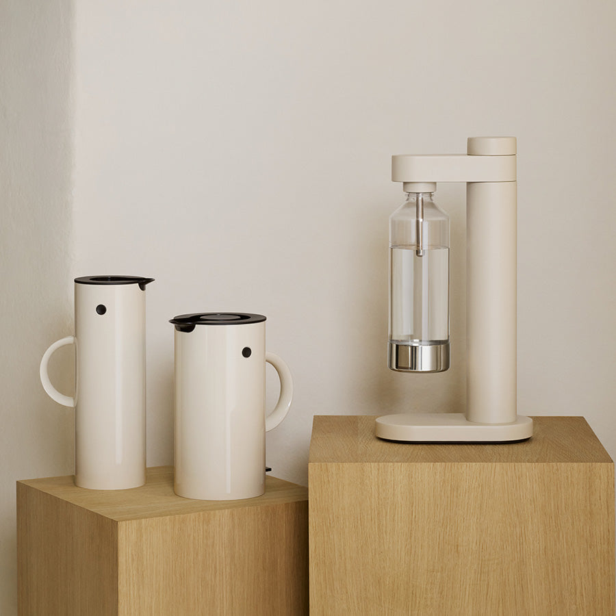 To Stelton EM77 termokanner i sandfarge plassert ved siden av en stilren vannkarafle, illustrerer moderne nordisk design og funksjonalitet.