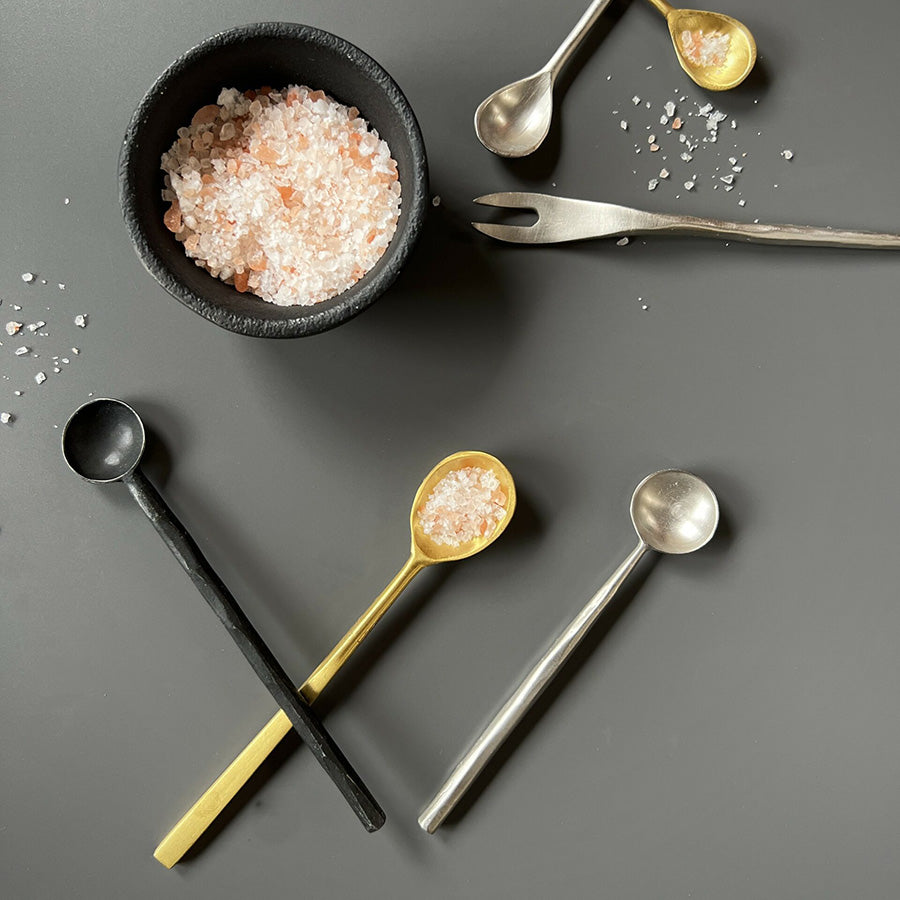 Tre forskjellige serveringsskjeer i forskjellige materialer og farger, inkludert sort, metallisk gull og rustfritt stål, brukt til å servere salt.