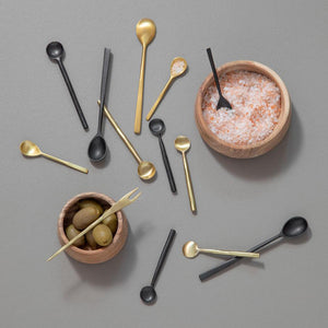 Variert utvalg av serveringsskjeer inkludert messing og sorte varianter, samlet rundt skåler med oliven og salt.