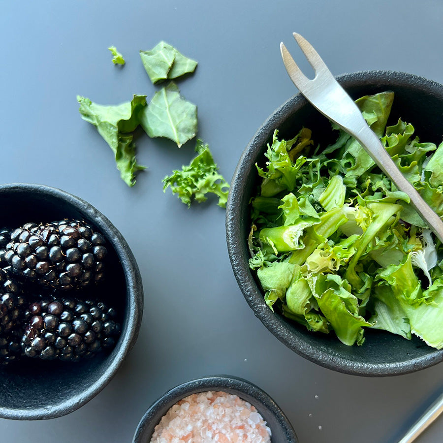 Serveringsgaffel i sølv med grønn salat og bær i sorte skåler, illustrerer bruken av gaffel for servering av friske ingredienser.