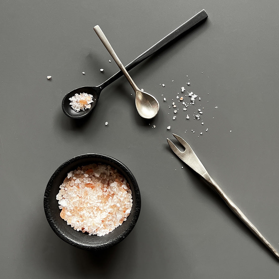 To stilige skjeer og en serveringsgaffel på en grå bakgrunn med havsalt i en svart skål, ideelt til tapas og delikate retter.