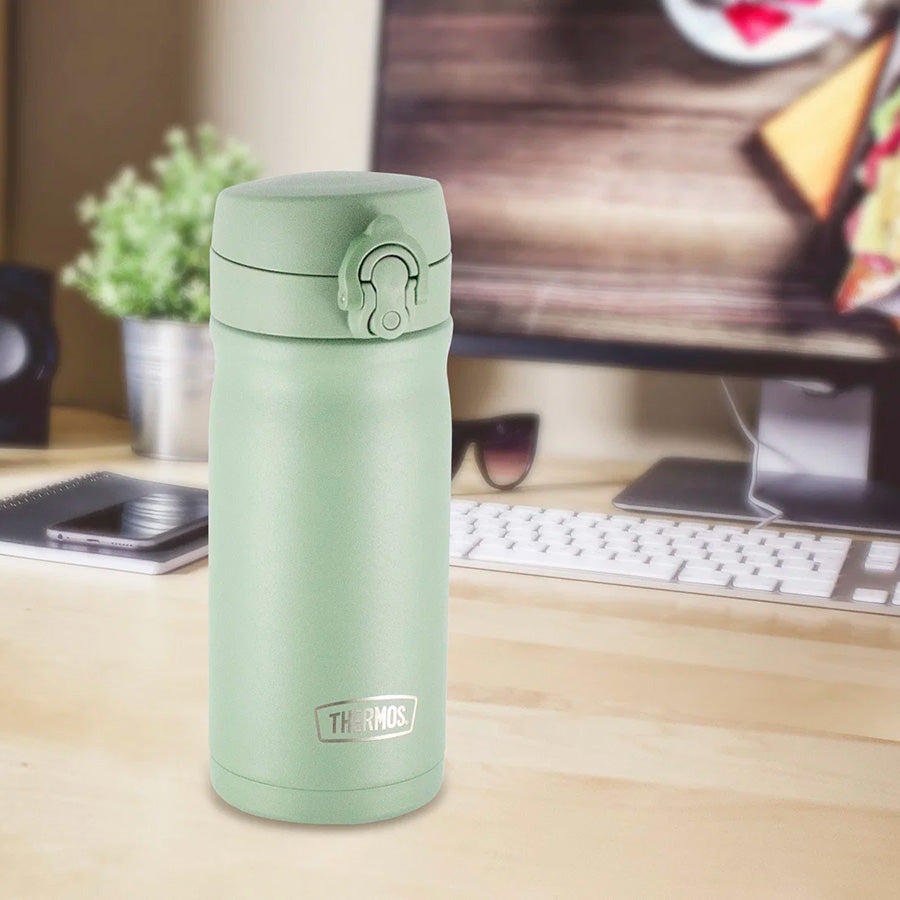 Thermos JMY termokopp i dusty green, plassert på et skrivebord med datamaskin, designet for elegant drikking.