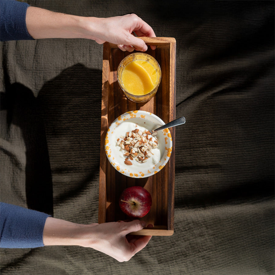 Hender som bærer Zassenhaus serveringsbrett med yoghurt, appelsinjuice og et eple, illustrerer brettets allsidighet.